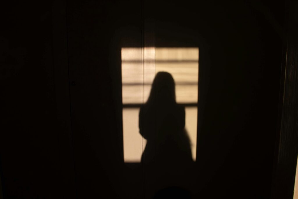 Silhouette of a woman standing near a window in dim light representing the social causes of sexual violence and silence in society